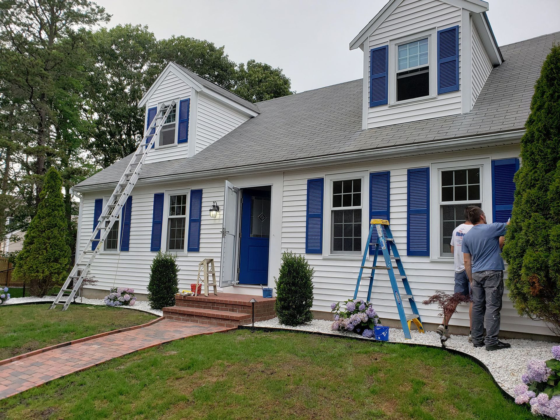 Workers painting a white house with blue shutters and trim, with ladders and tools in the yard.