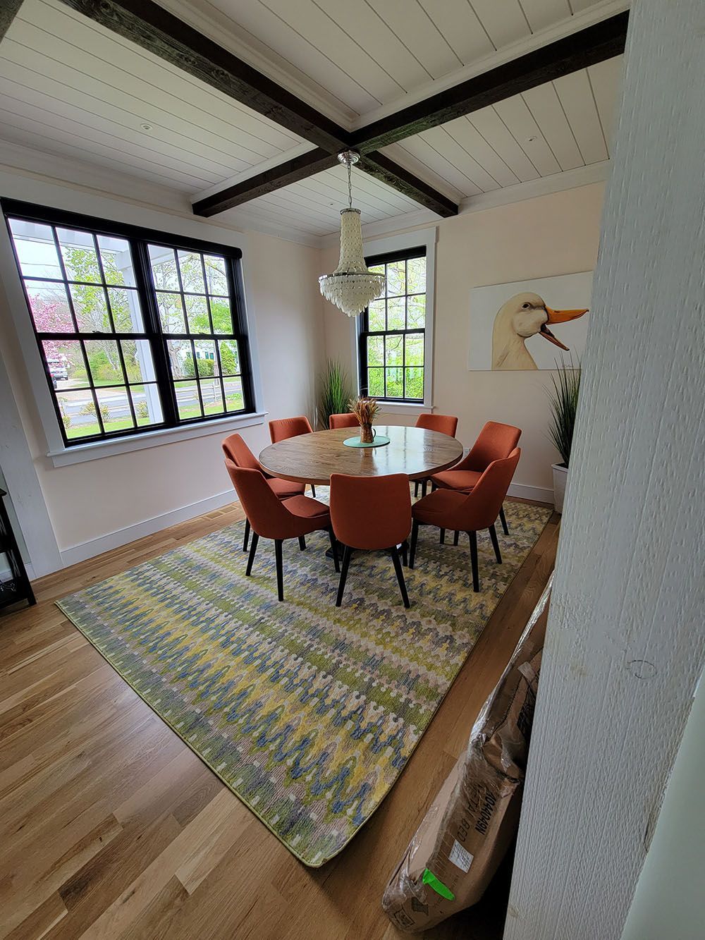 Bright dining room with round table, orange chairs, patterned rug, large windows, and a hanging light fixture.