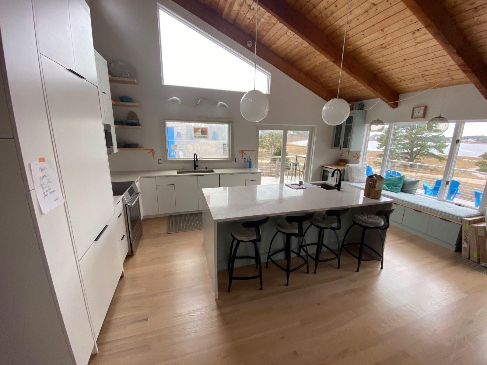 Modern kitchen with island, white cabinets, wood ceiling, and large windows overlooking trees and a pool.