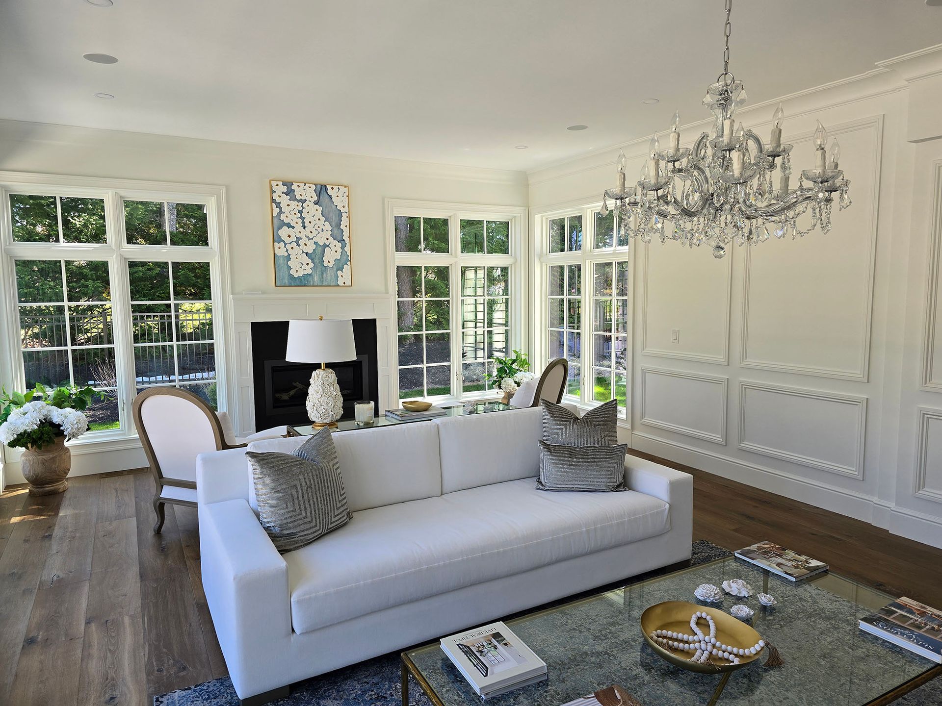 Elegant white living room with chandelier, fireplace, large windows, and gray sofa.