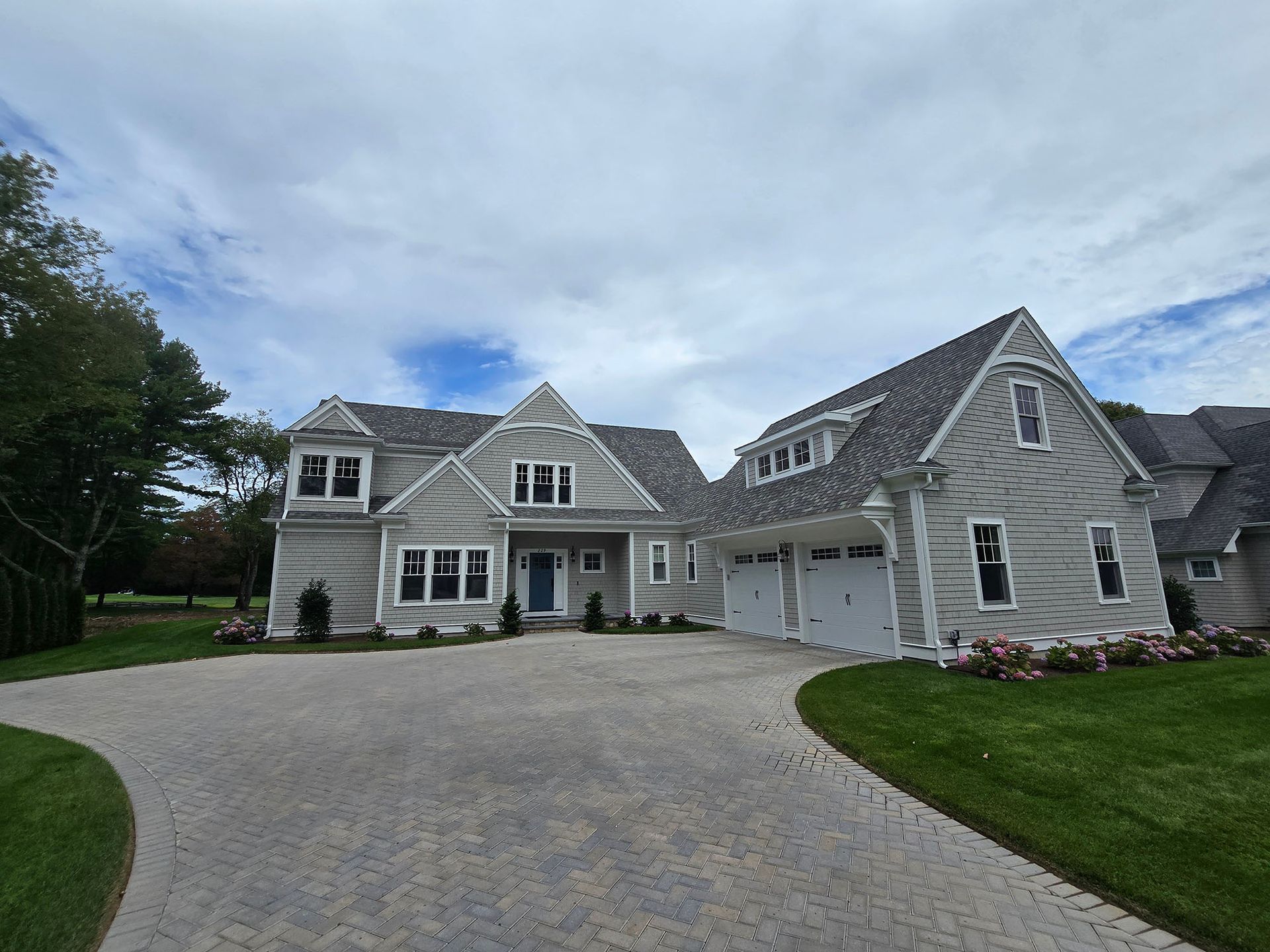 Large gray suburban house with a wide driveway, green lawn, and cloudy sky