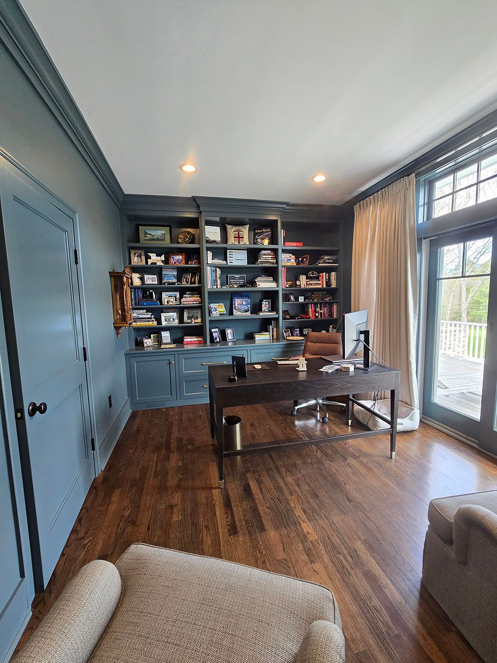 Home office with blue built-in shelves, a desk, hardwood floors, and large windows.