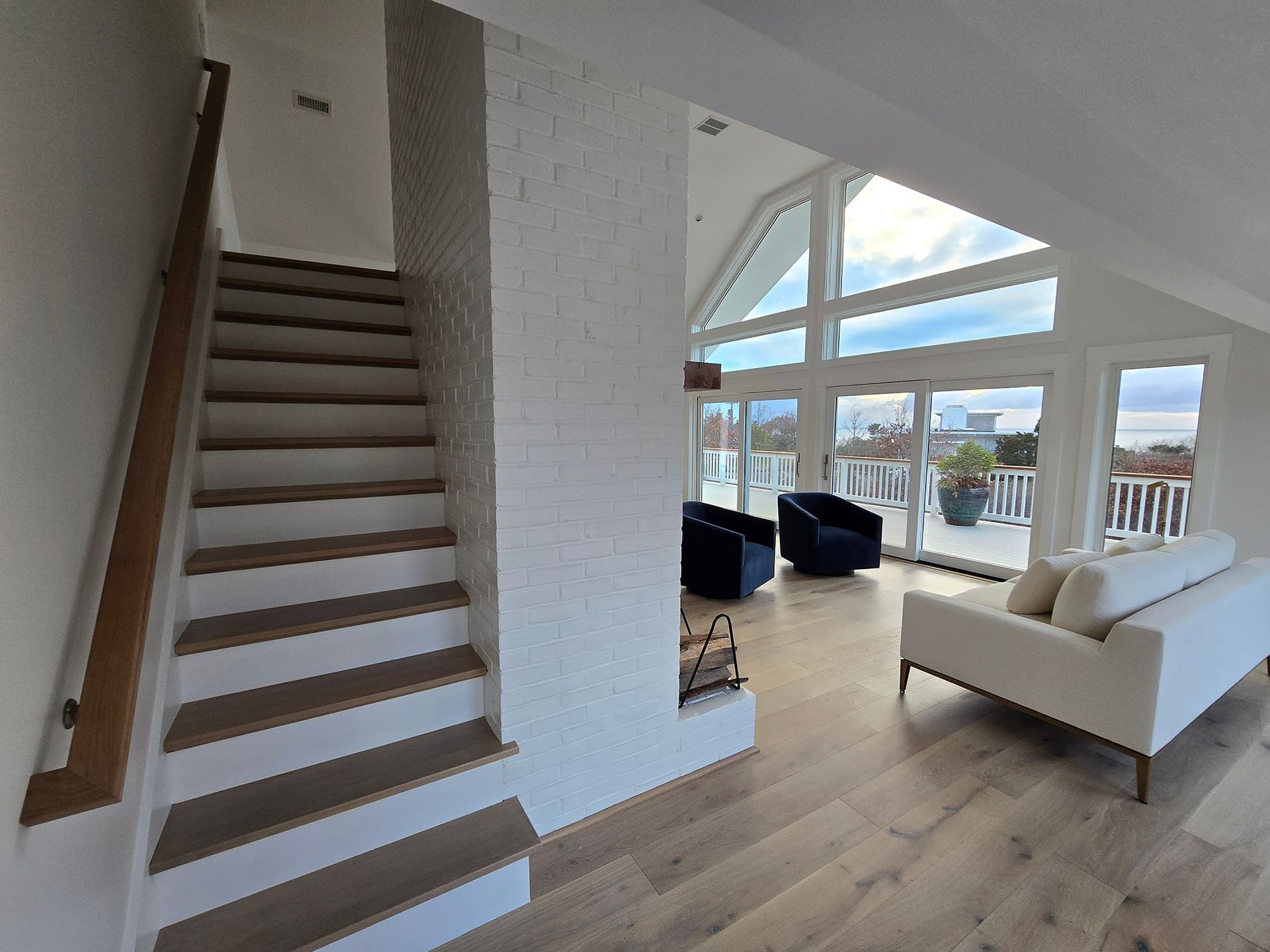 Bright modern living room with staircase, white sofa, black chairs, and large windows overlooking water.