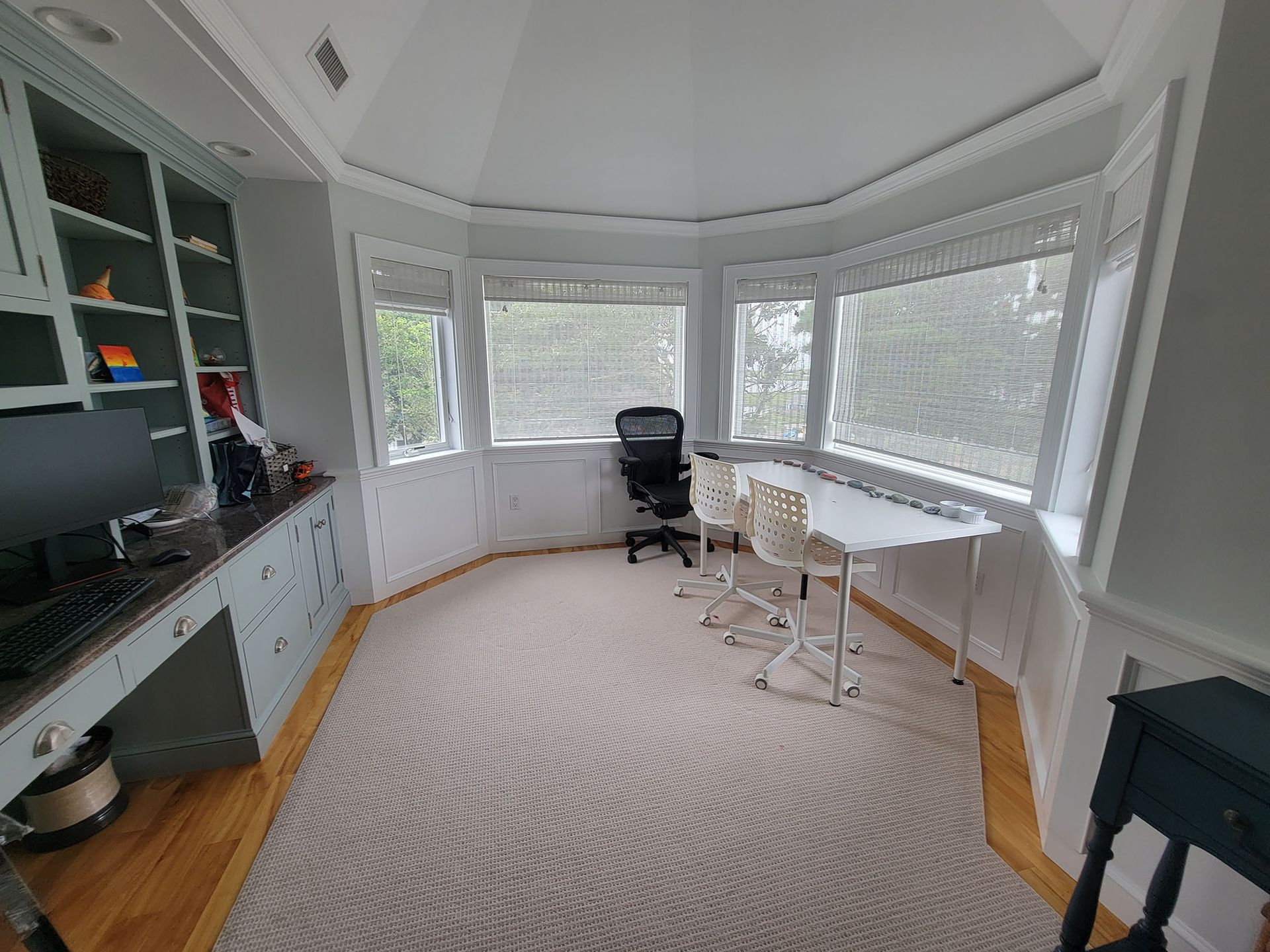 Bright sunroom office with bay windows, desk, chair, shelves, and white furniture over a patterned rug.