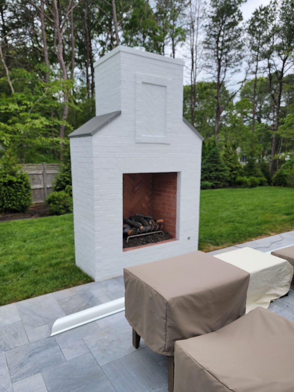 White outdoor brick fireplace by a patio with covered chairs, surrounded by a green lawn and trees