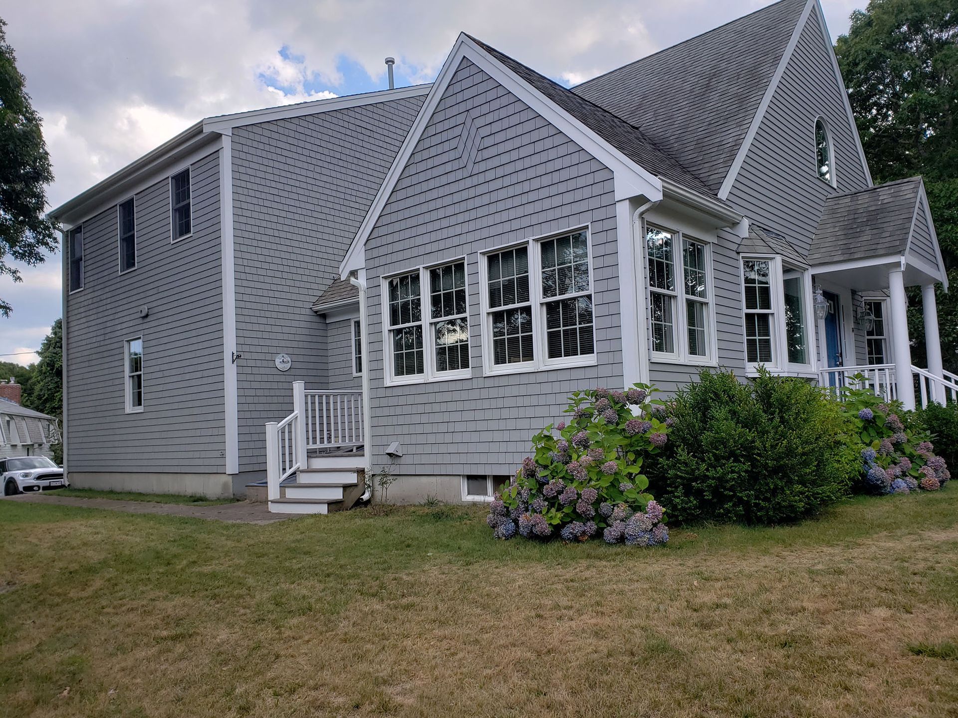 Gray shingled house with white trim, dormer windows, and a small front porch beside a lawn and shrubs