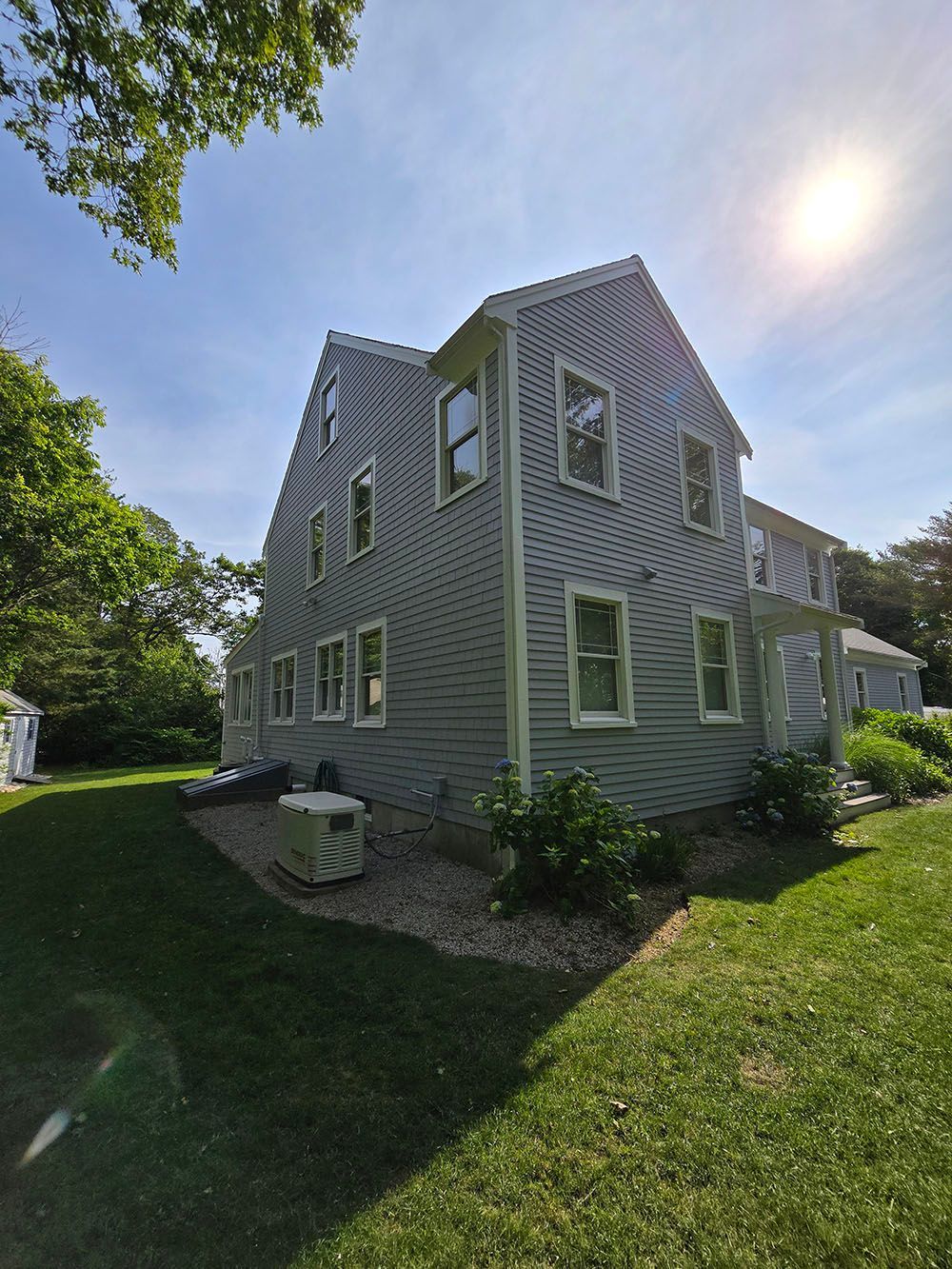 Gray shingled house with white trim on a sunny lawn, seen from the side with trees overhead.