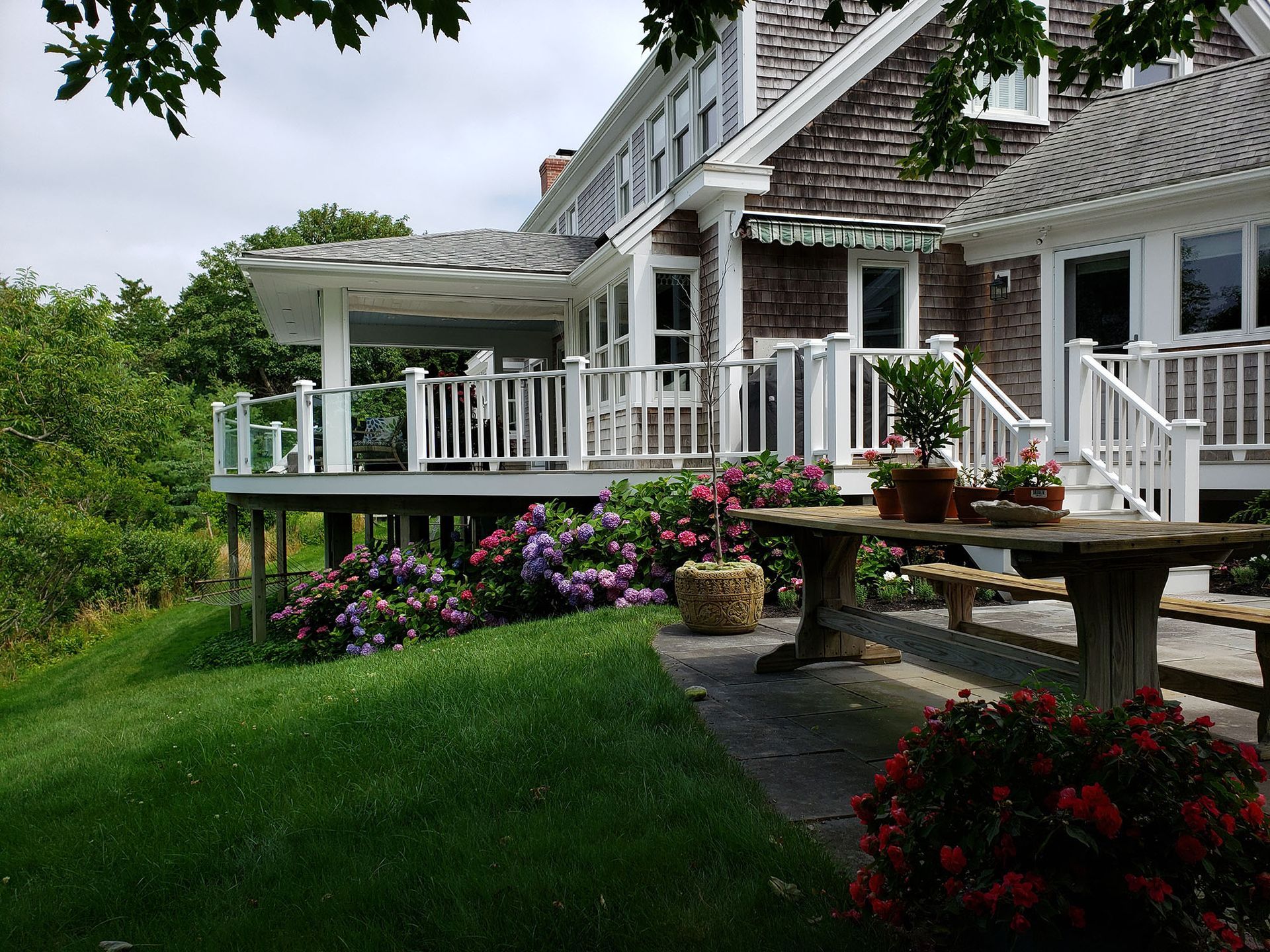 White house with wraparound deck, white railing, and colorful flowers beside a green lawn