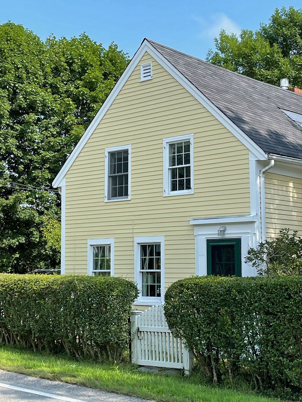 Yellow cottage with white trim and green door behind a hedge on a sunny day