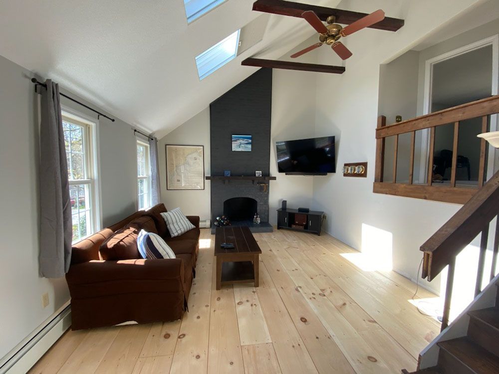 Bright living room with wooden floor, brown sofa, wall TV, skylight, and staircase.