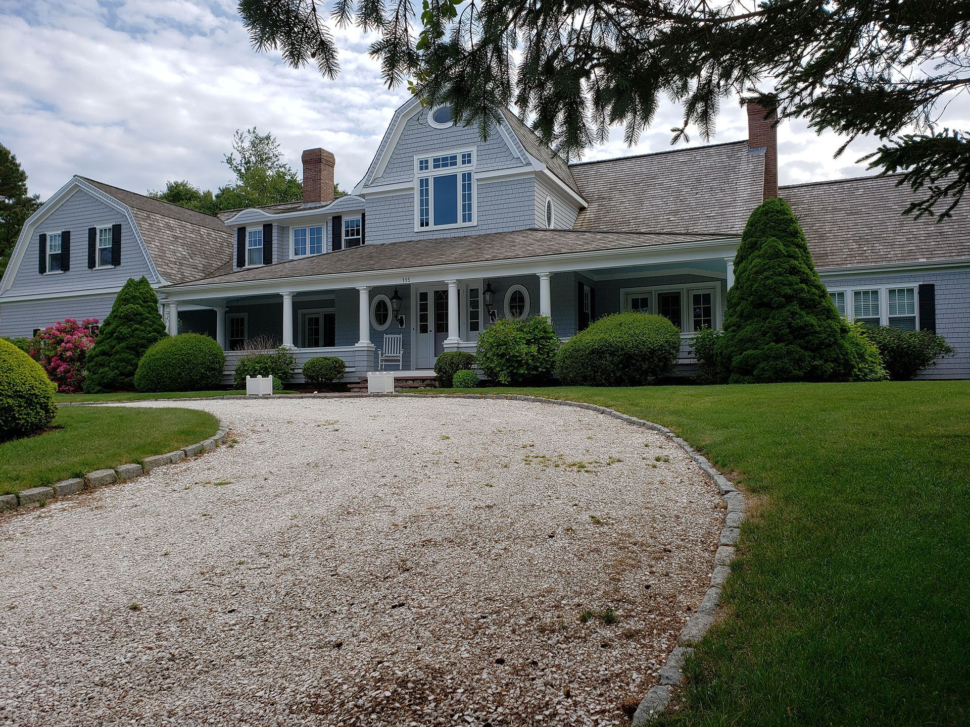 Large gray shingle house with a curved gravel driveway, white porch, and manicured lawn