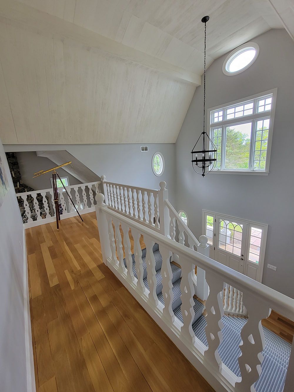 Bright upstairs hallway with white railings, wood floor, large windows, and a chandelier.