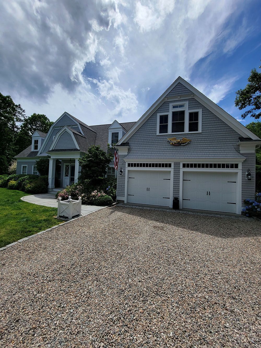White house with gray shutters, gravel driveway, flower boxes, and a double garage under a blue sky