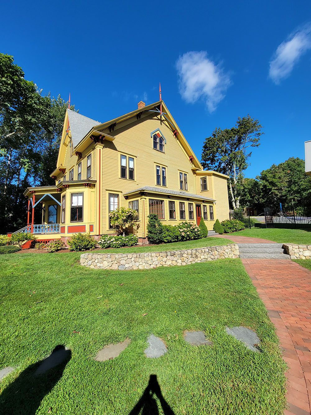 Yellow Victorian-style house with steep gables and wraparound porch on a sunny lawn under a blue sky