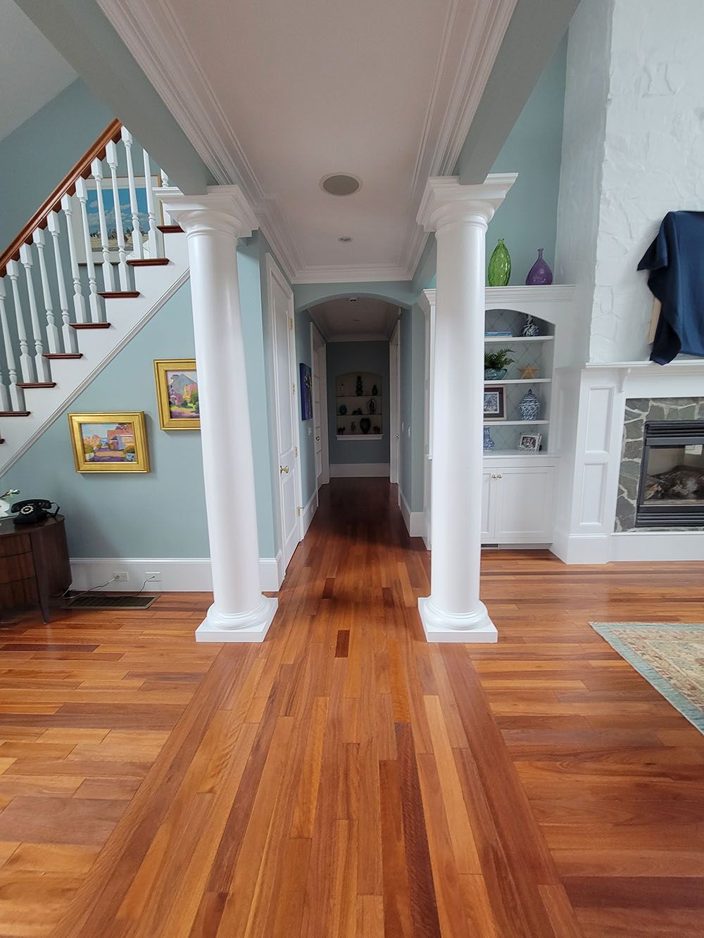 Hallway with polished hardwood floors, white columns, staircase, and built-in shelves in a bright home.