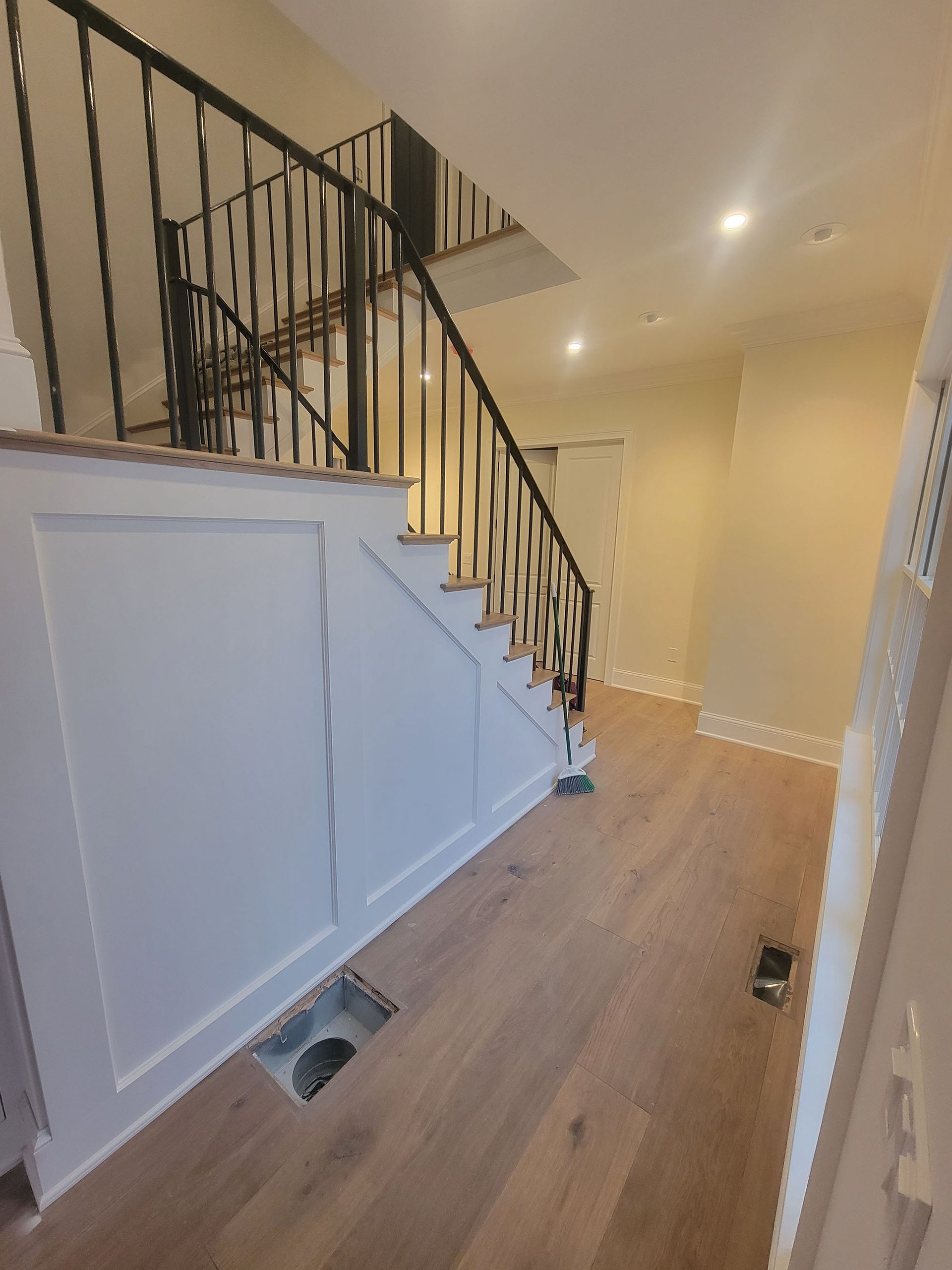 Hallway with light wood floors, white paneled walls, and a black staircase railing under warm ceiling lights