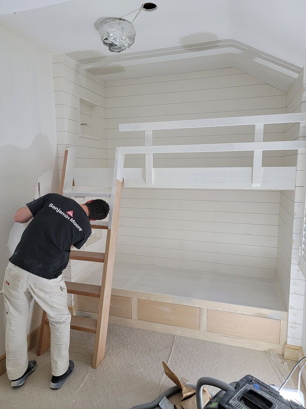 Worker assembling a white bunk bed in a bright, empty bedroom with a ladder and tools on the floor