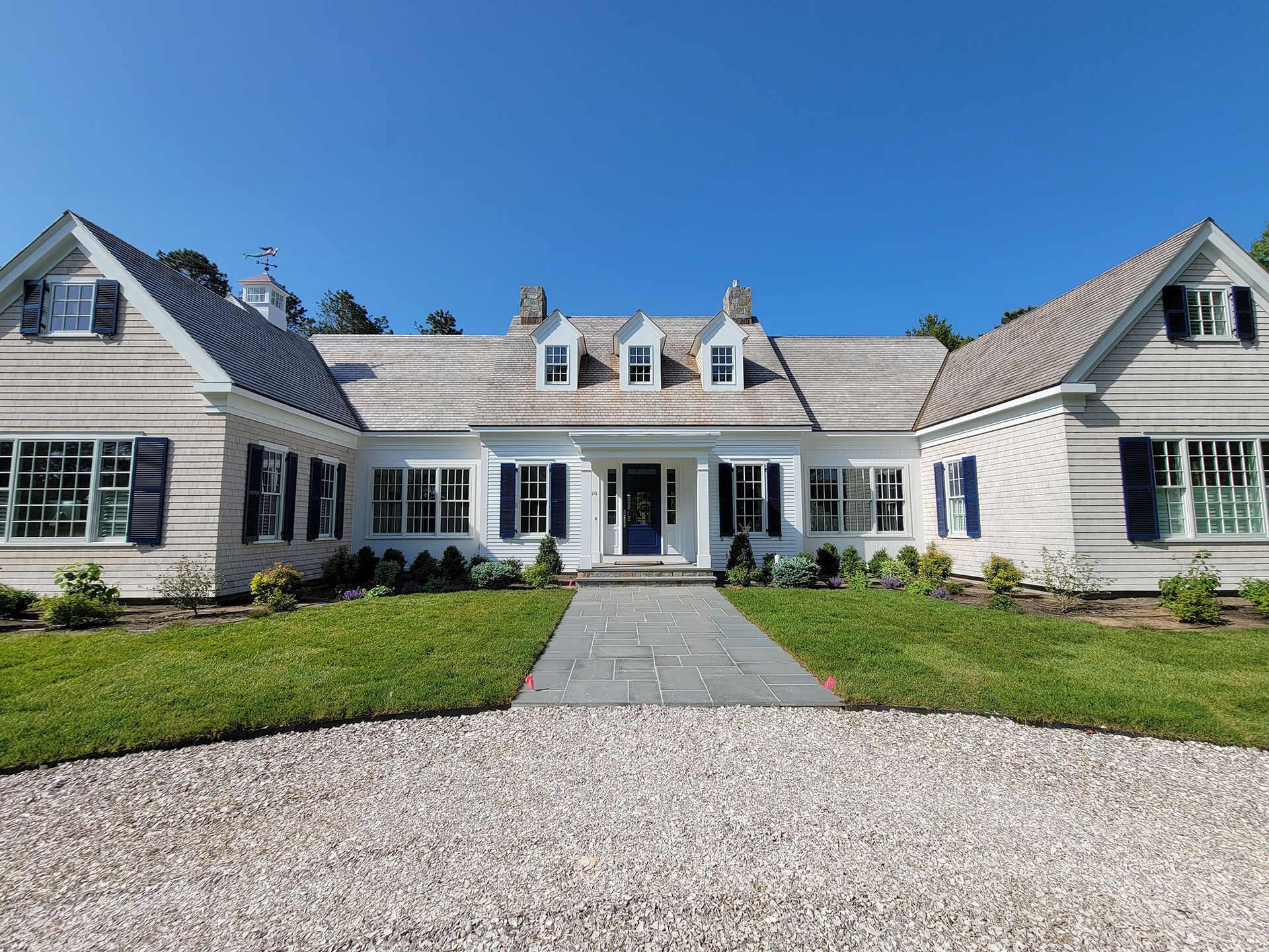 Large white house with a gray shingled roof, central walkway, and gravel driveway under a clear blue sky