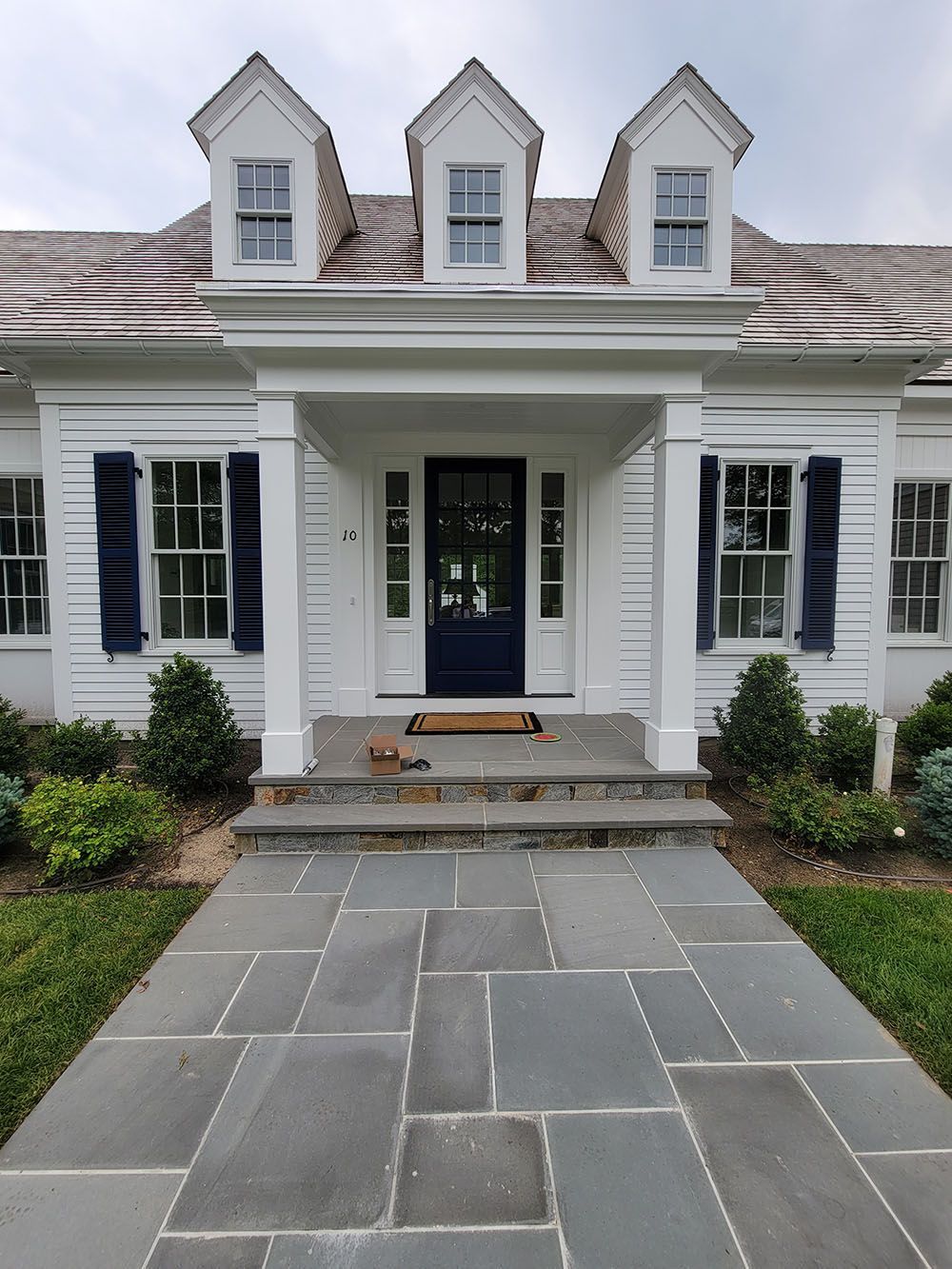White house entrance with black shutters, stone walkway, and three dormer windows