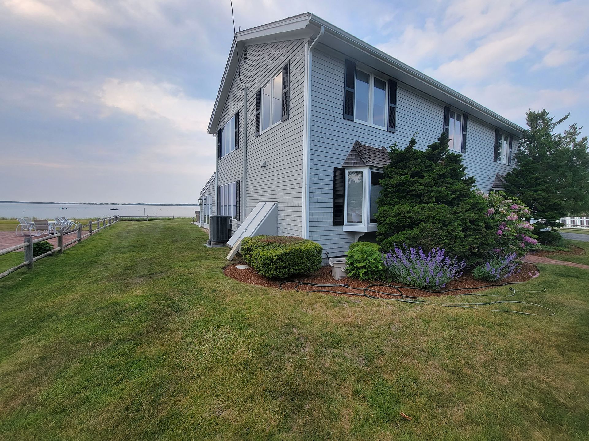 Two-story gray house with white trim on a grassy hill by the water at sunset