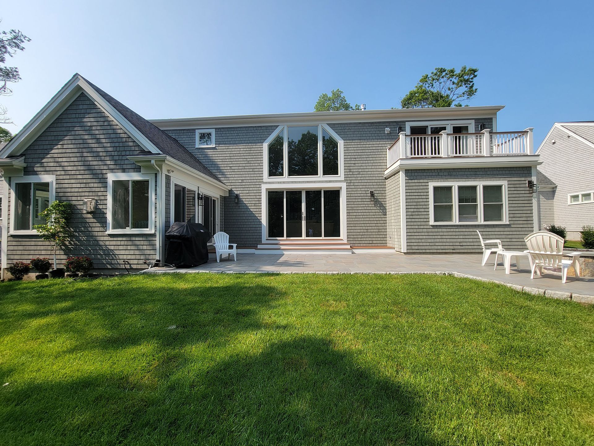 Backyard view of a gray-shingled house with large windows, patio seating, and a green lawn