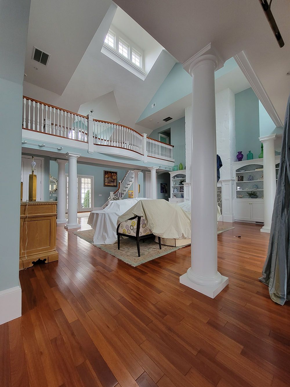 Bright airy living room with hardwood floors, tall white columns, skylight, and covered furniture in the center.