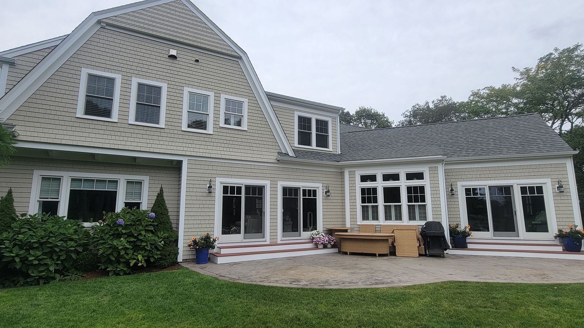 Gray shingled house with white trim, multiple windows, and a patio with outdoor furniture under cloudy skies