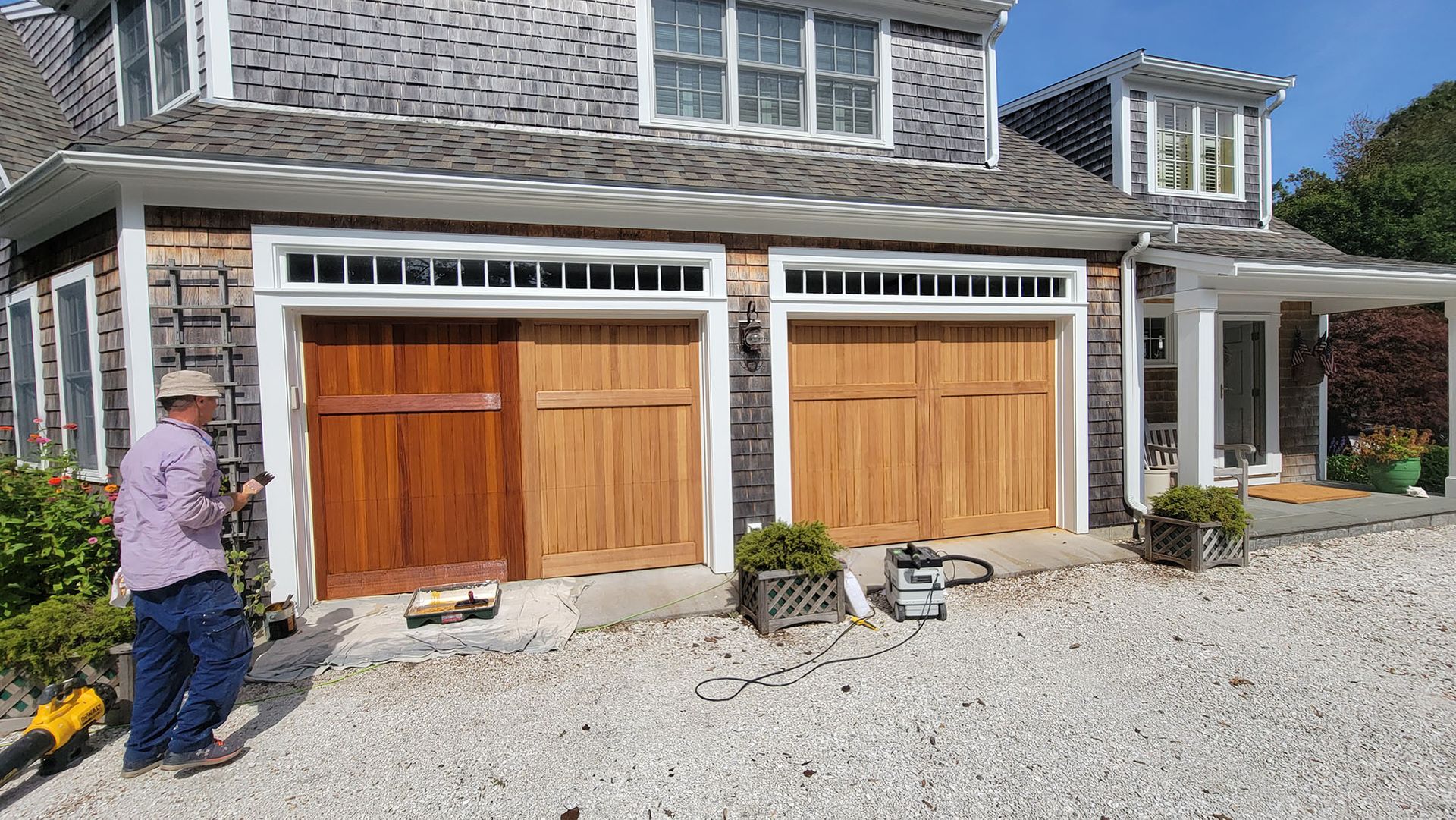 Two wooden garage doors on a stone house, with a person standing on the left and tools on the driveway.
