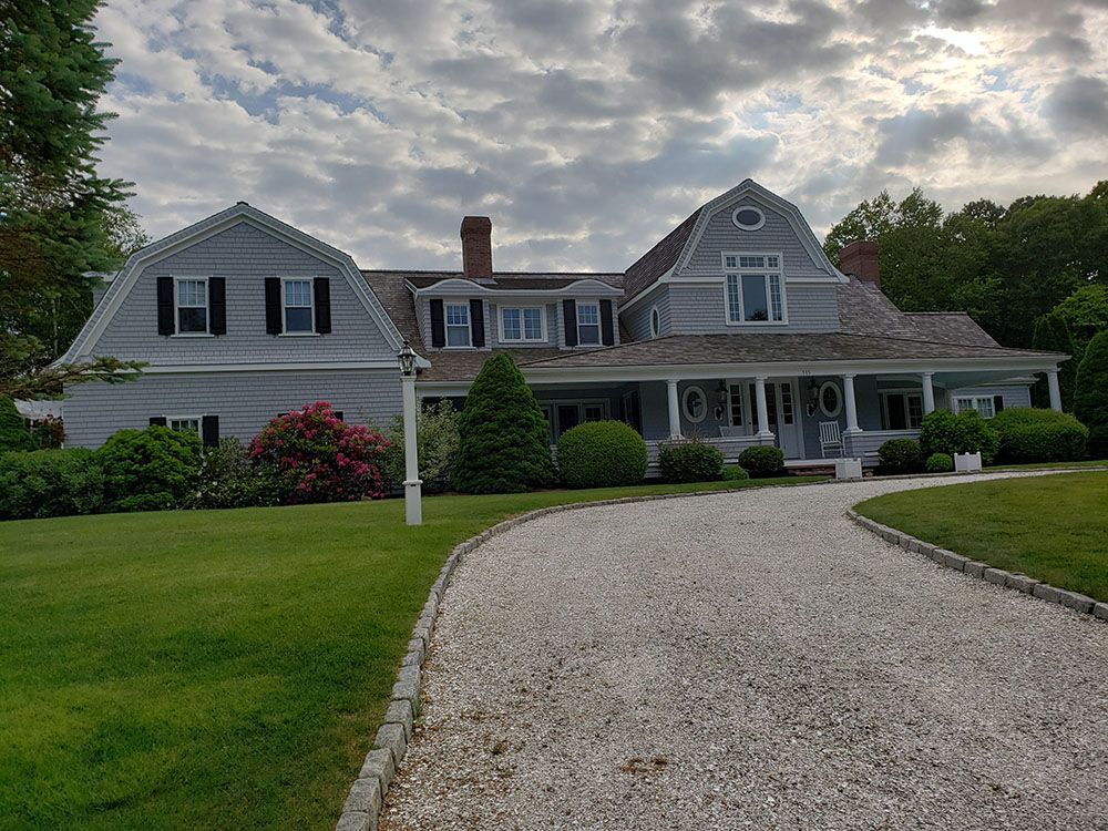 Large gray suburban house with a curved gravel driveway and manicured lawn under a cloudy sky