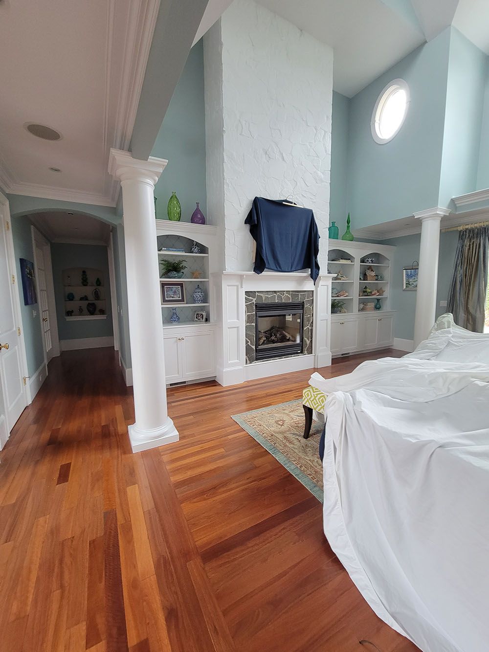 Bright bedroom with high ceiling, white fireplace, hardwood floors, and a bed with white bedding.