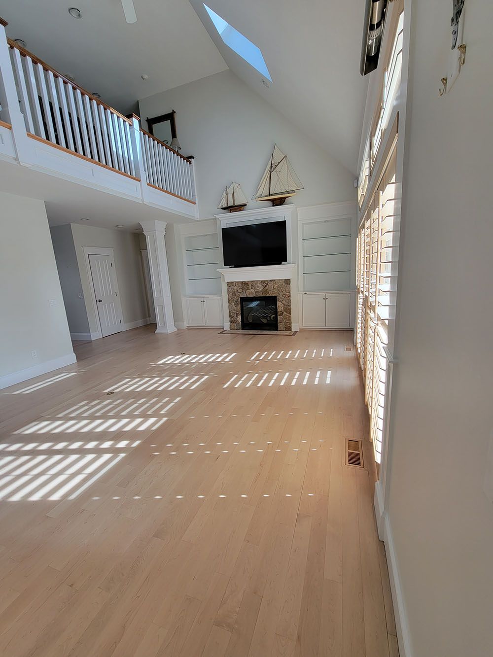 Bright two-story living room with white walls, loft balcony, fireplace, and sunlight streaming through tall windows