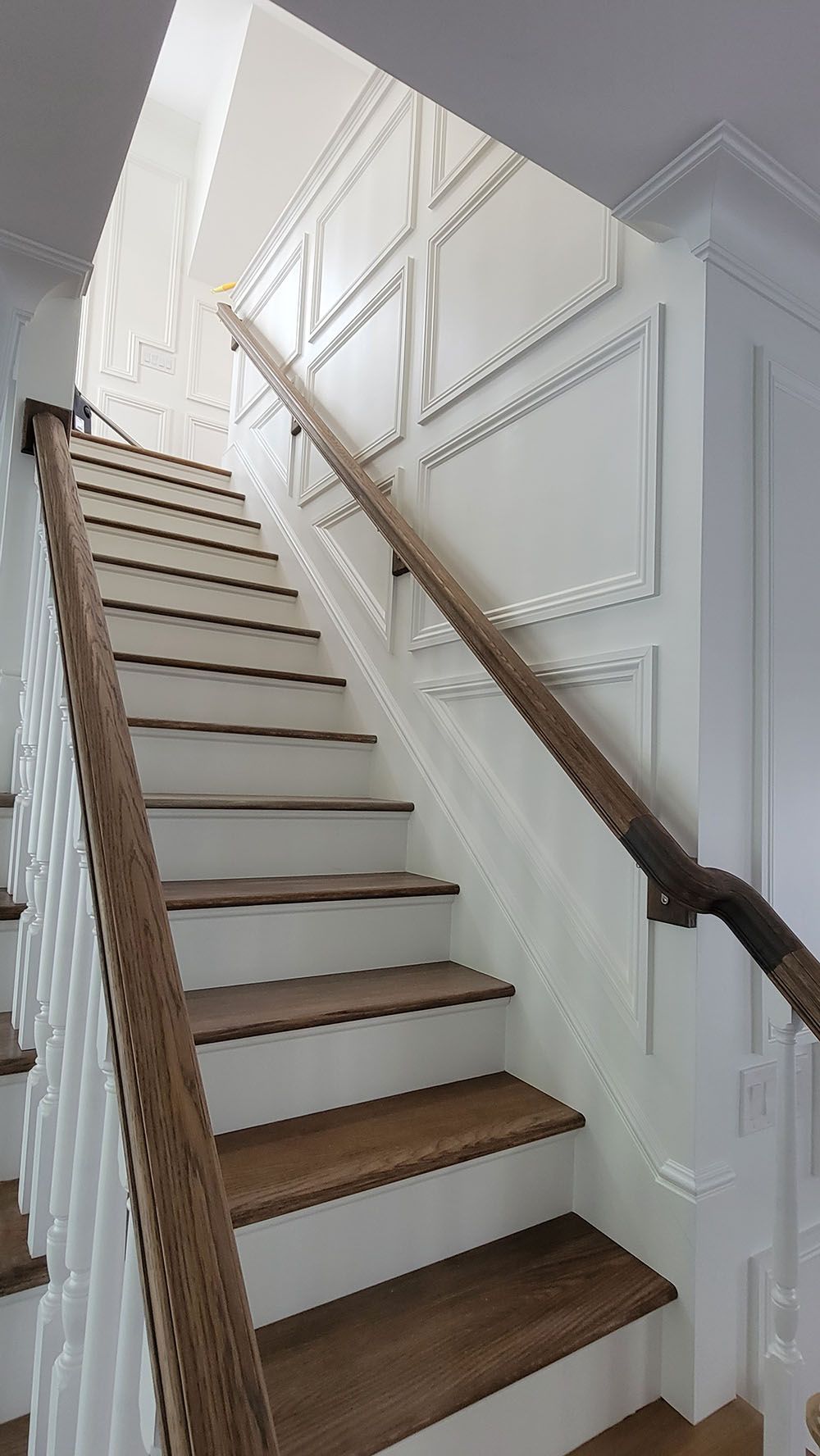 Narrow staircase with white paneled walls, wood treads, and a dark handrail in a bright interior.