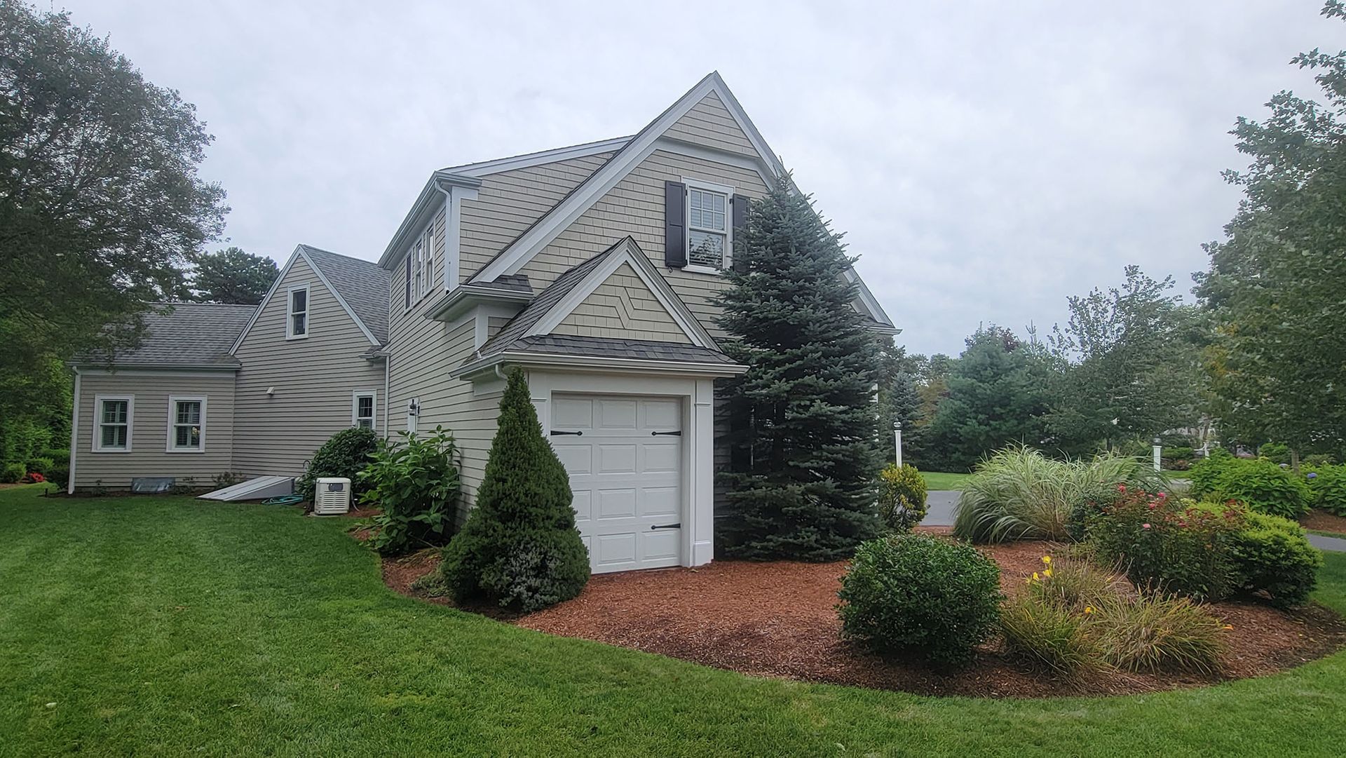 Large gray house with white garage, landscaped garden, and green lawn under a cloudy sky