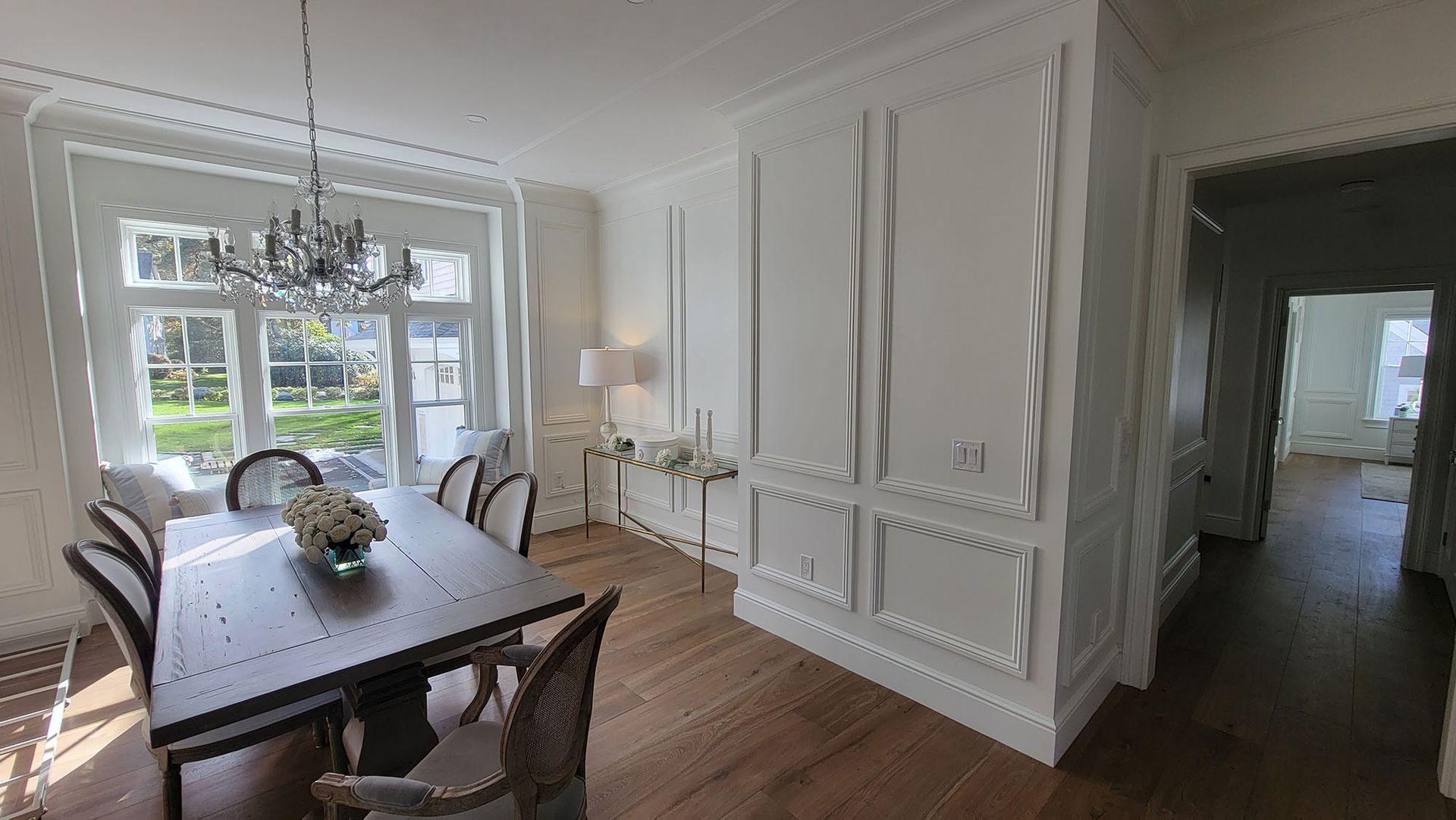 Elegant dining room with long table, woven chairs, white paneled walls, and a chandelier by bright windows