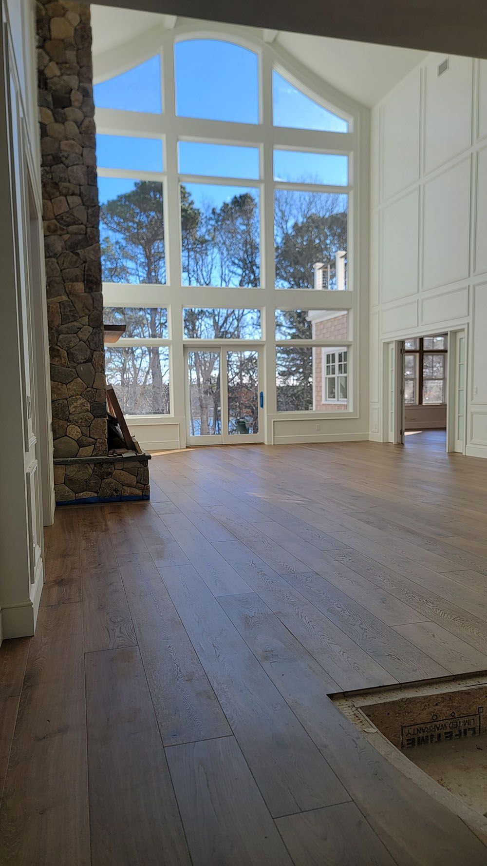 Bright living room with tall arched windows, wood floors, and a stone fireplace, overlooking trees outside