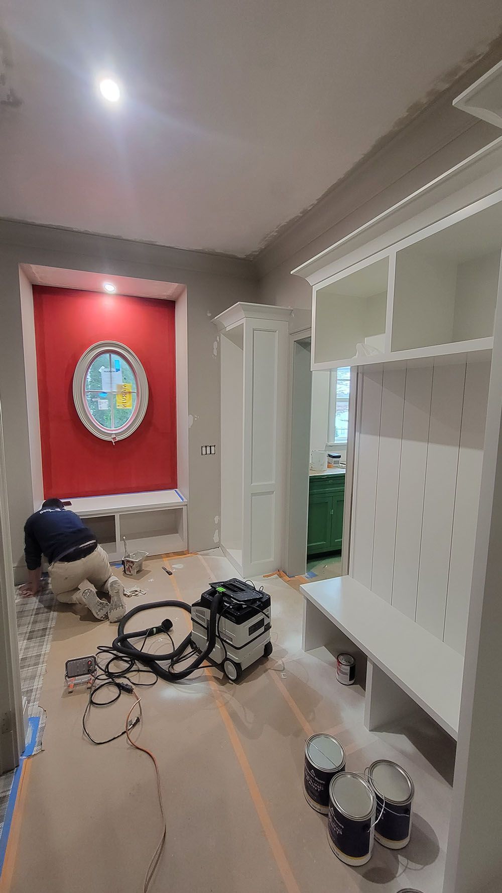 A room under renovation with tools, cables, and paint cans; a worker kneels near a red-covered window.