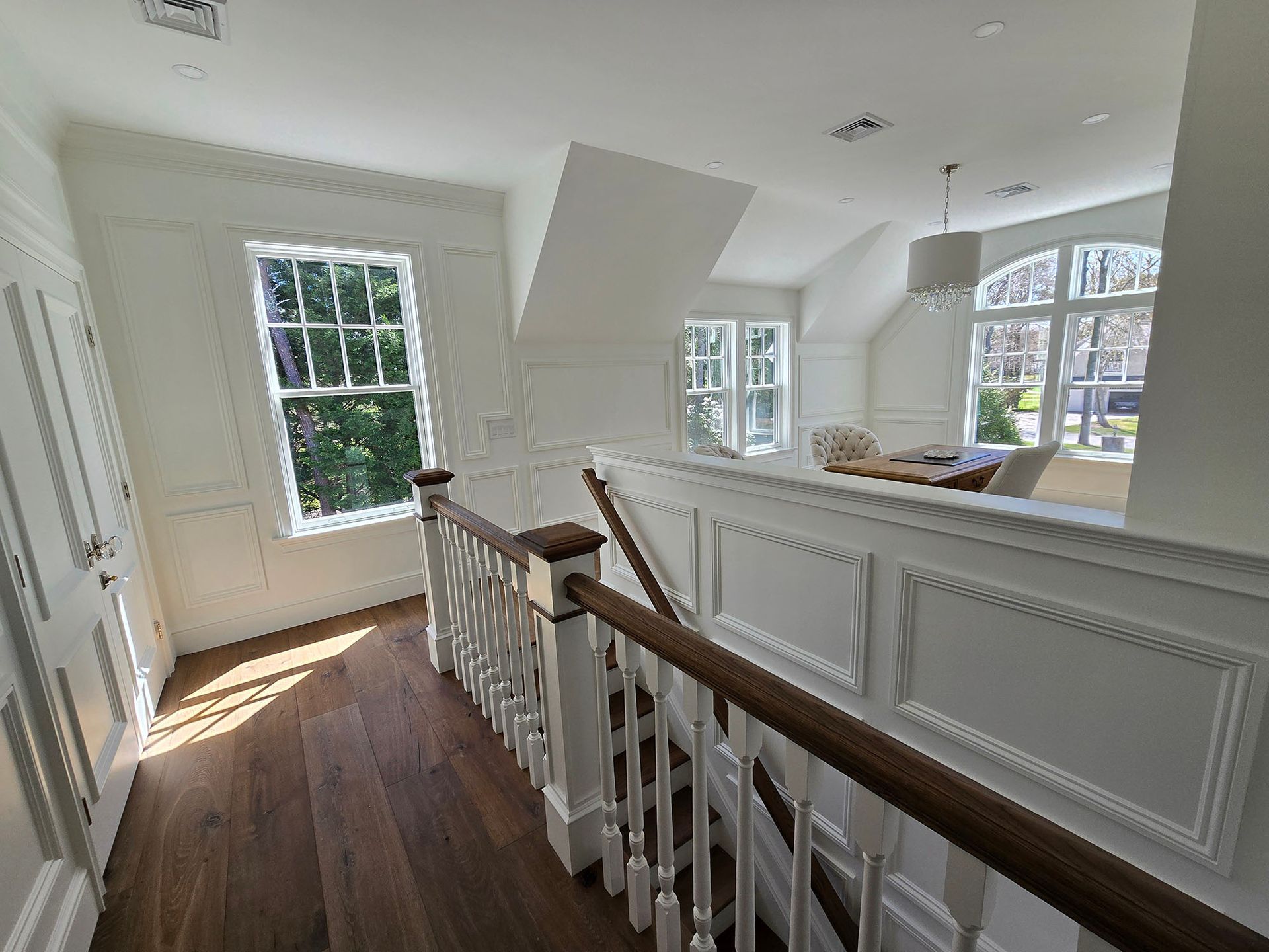 Bright hallway with white walls, dark wood floors, staircase railing, and large windows letting in daylight