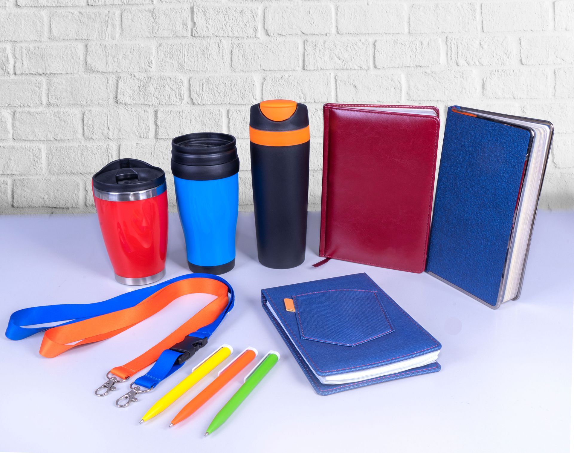 Promotional items: red and blue mugs, pens, lanyards, and notebooks arranged against a white brick wall.