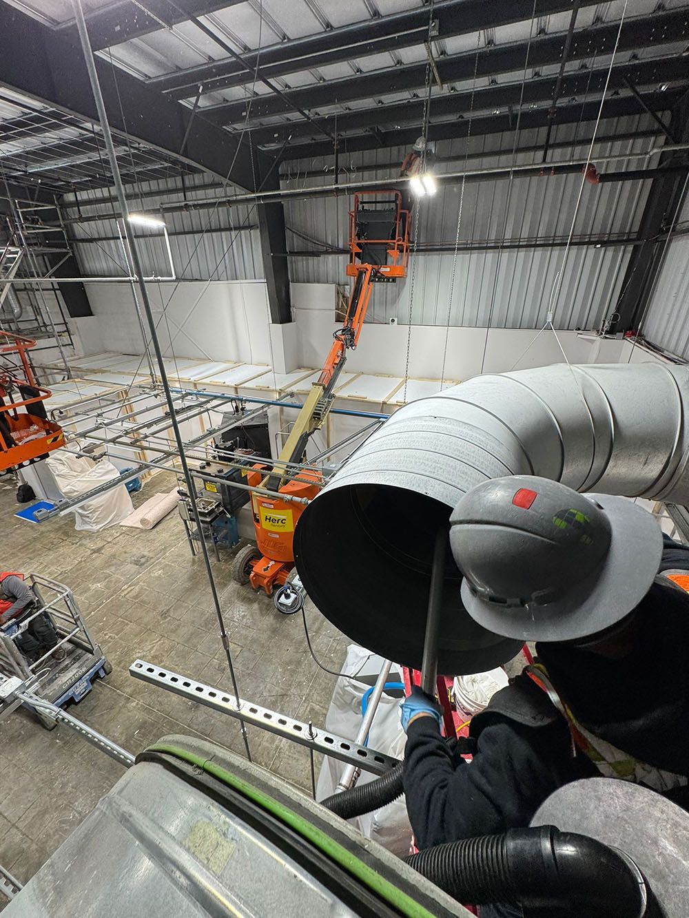 Worker on an orange lift inspecting large gray HVAC ducts inside an industrial warehouse