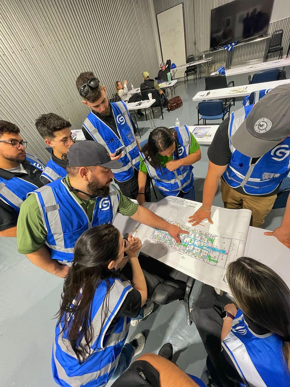 Group of volunteers in blue vests huddle around a table, planning over papers in a conference room.