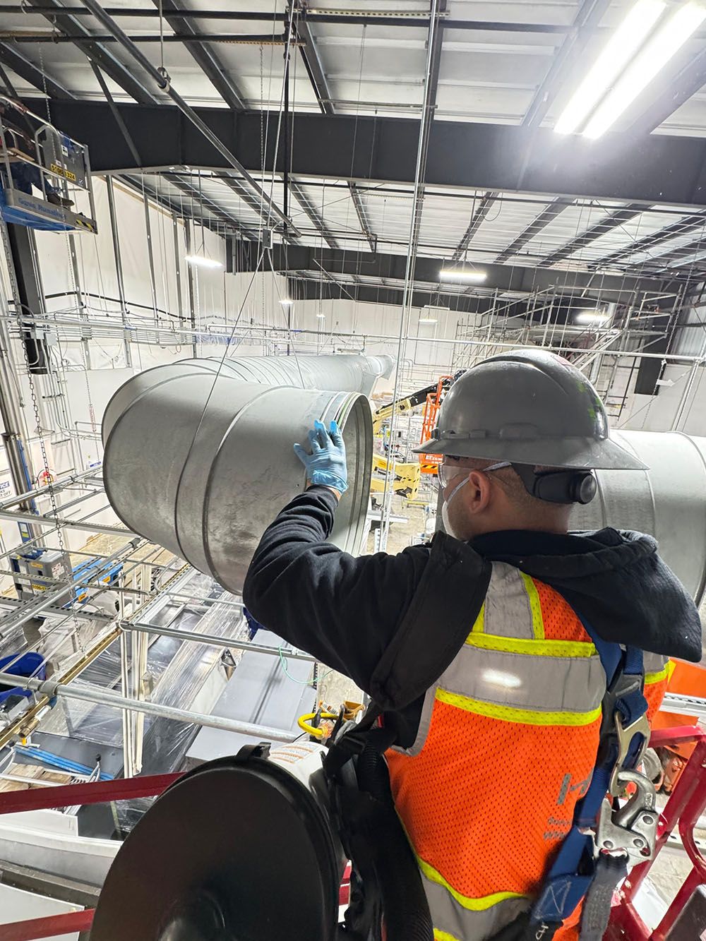 Worker in hard hat and orange vest operating large industrial equipment inside a metal-framed facility