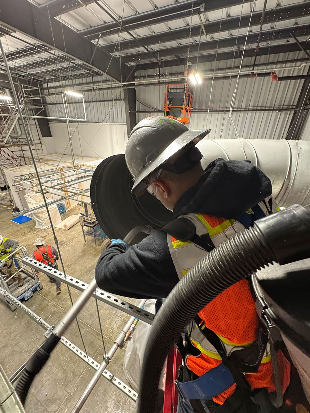 Worker in hard hat and safety vest climbing a metal structure inside a warehouse ceiling.