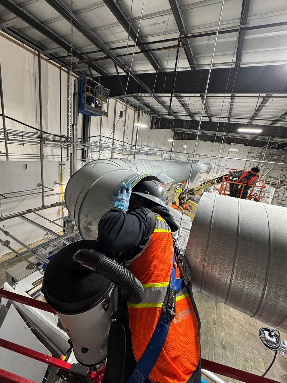 Worker in orange safety vest welding large metal pipes inside an industrial workshop