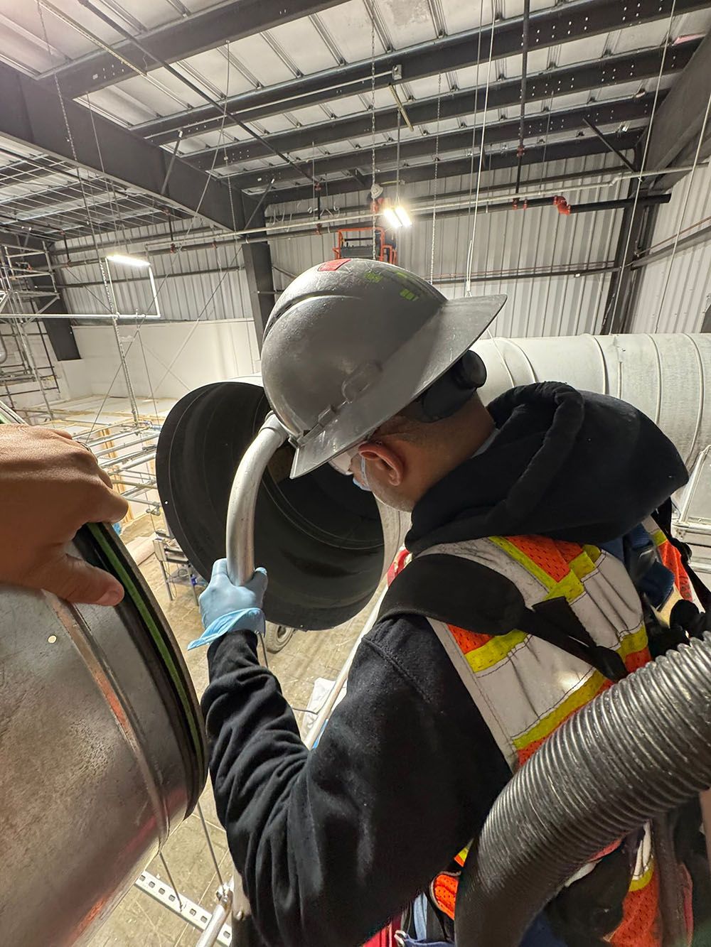 Worker in hard hat and gloves inspecting a large metal cylinder inside an industrial warehouse.
