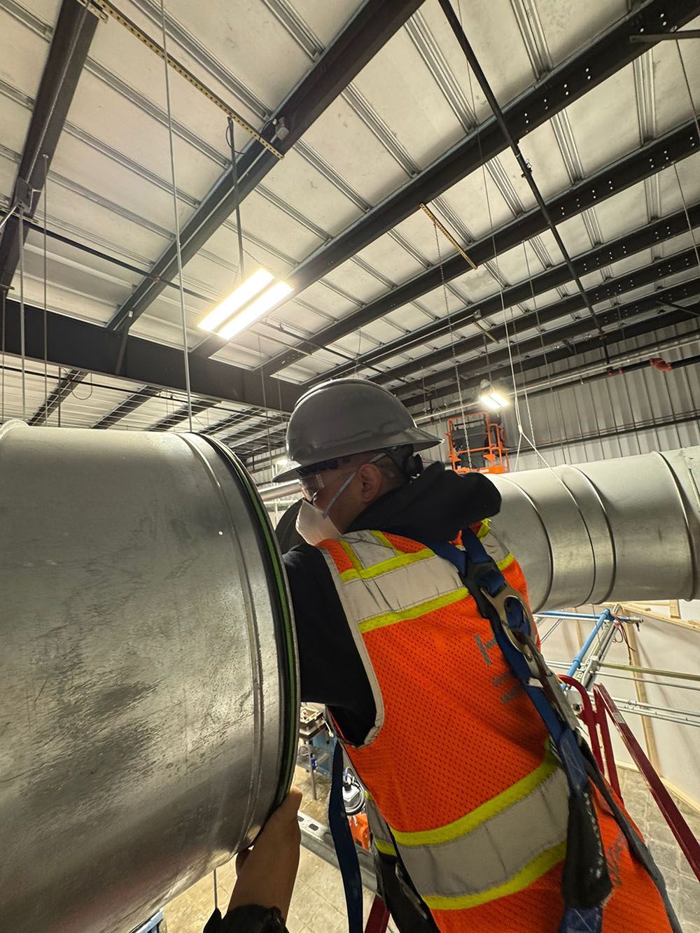 Worker in orange safety vest inspecting large metal ductwork inside an industrial warehouse.