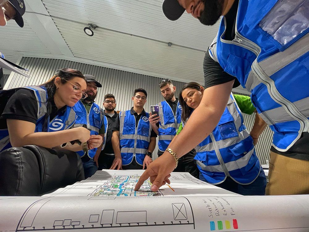 Group in blue vests gathers around a floor plan, pointing and discussing in a training room.