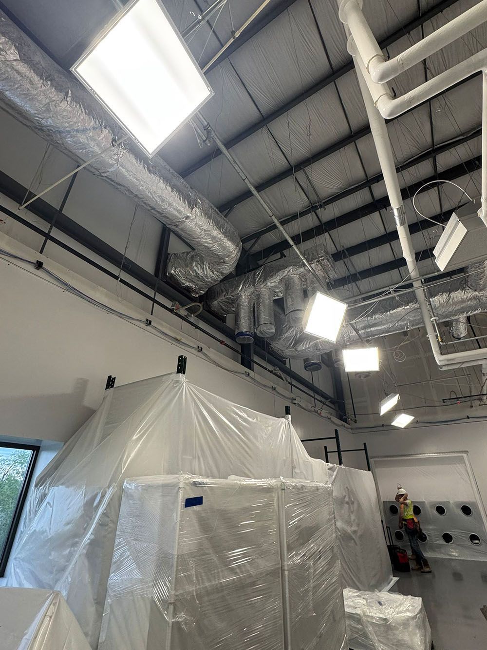 Industrial ceiling and plastic-covered equipment in a warehouse, with workers near a wall on the right.