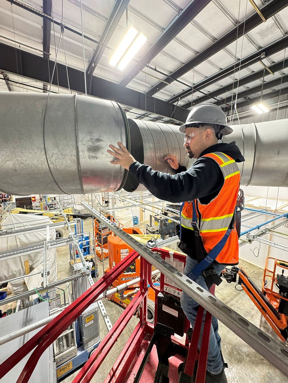 Worker in hard hat and safety vest handling a large metal pipe on a platform inside an industrial facility