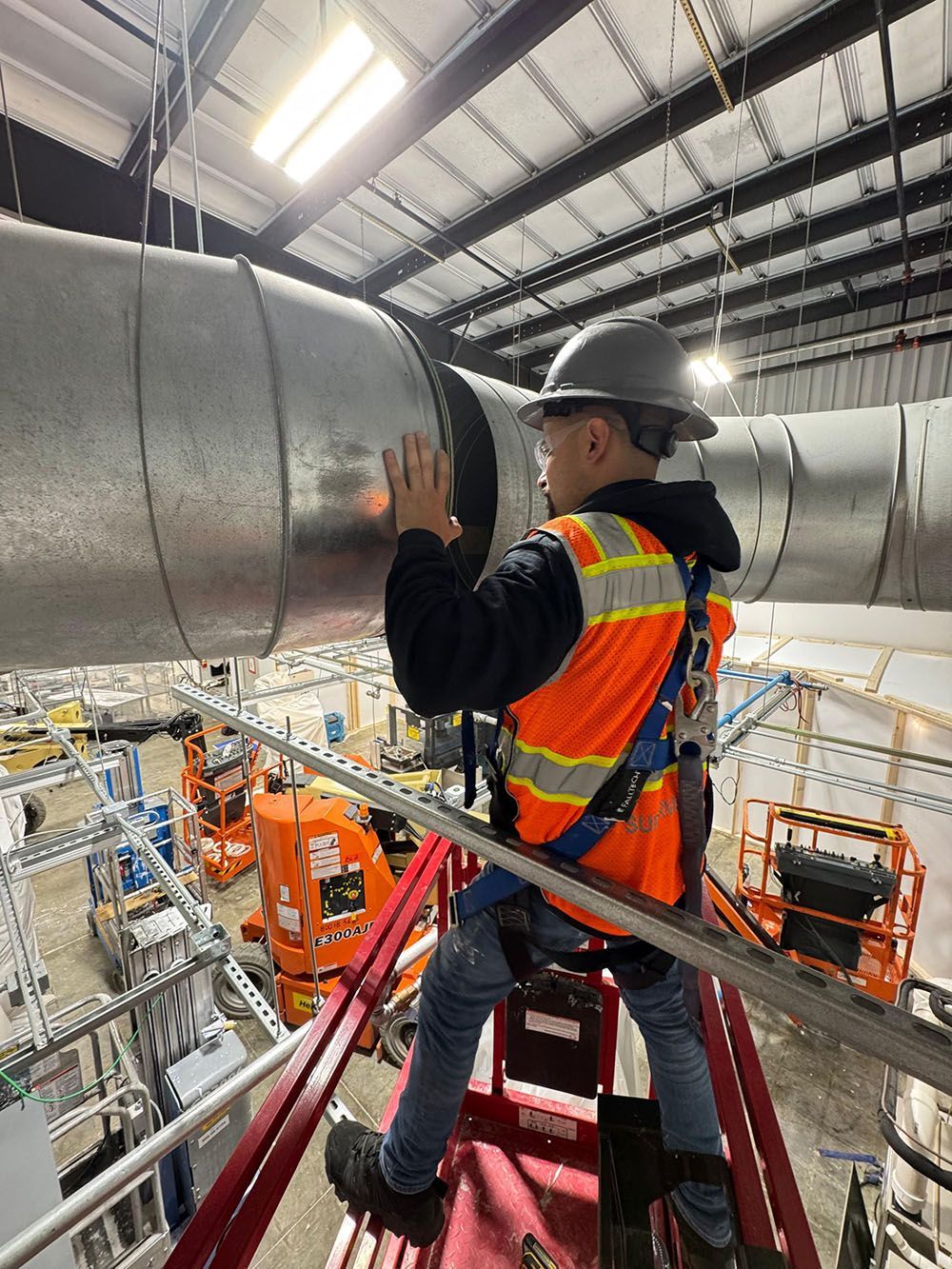 Worker in hard hat and safety vest inspecting large industrial ducting in a factory catwalk.