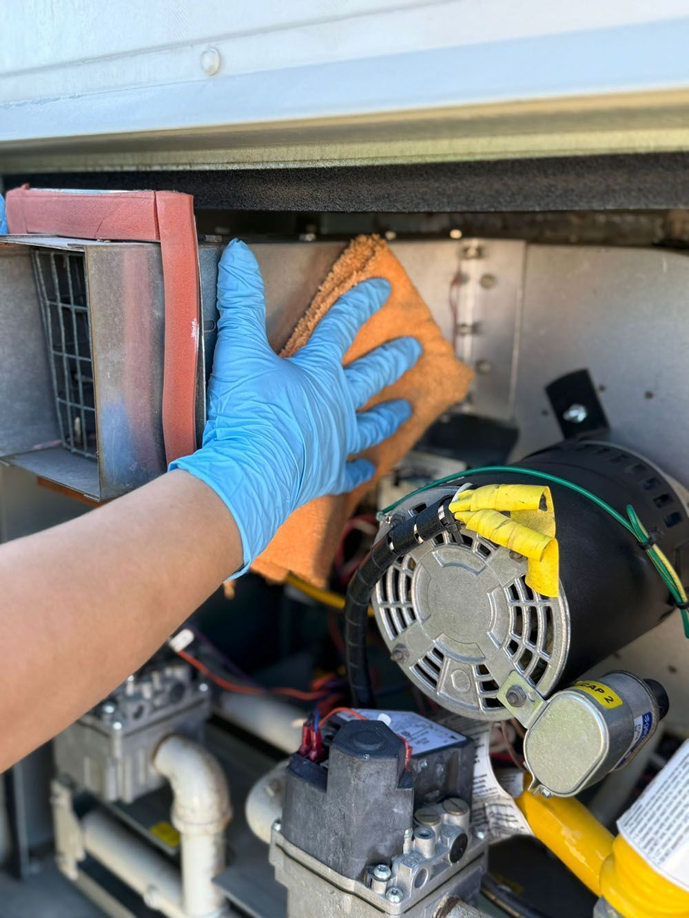 Blue-gloved hand cleaning an HVAC unit with an orange sponge in a cramped utility compartment